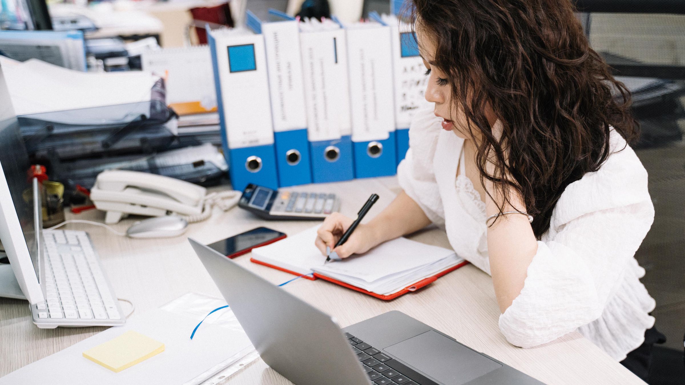 An office worker writes notes while using two laptops at a document-filled desk