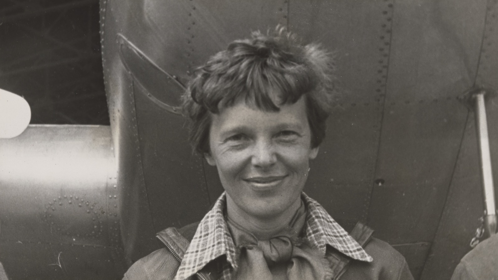 Black-and-white portrait, probably from March 1937, of Amelia Earhart smiling beneath the riveted nose of her Lockheed Model 10-E Electra.