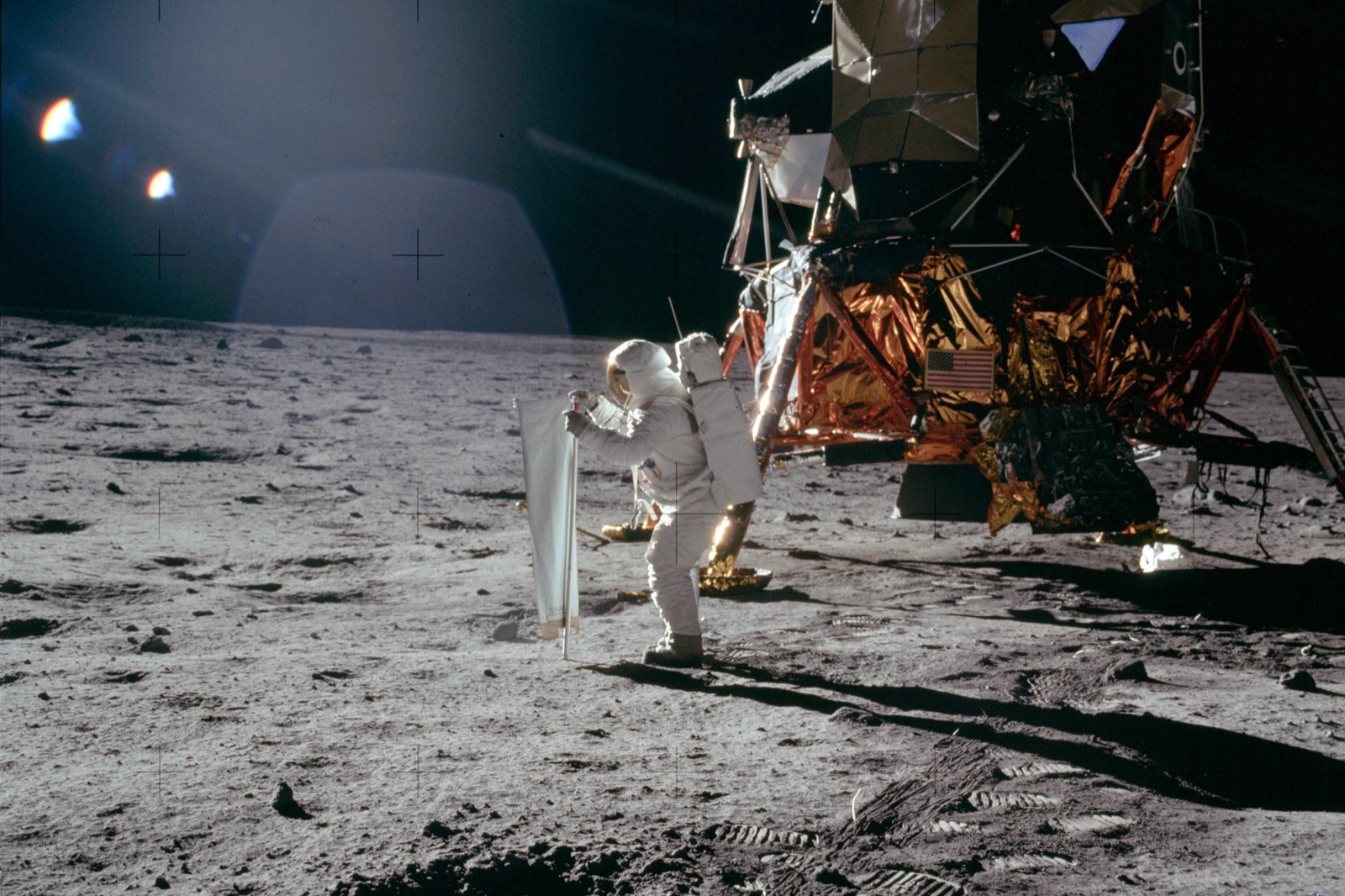 Buzz Aldrin stands beside the Apollo 11 lunar module while unfurling a bright rectangular sheet of aluminum foil on a pole
