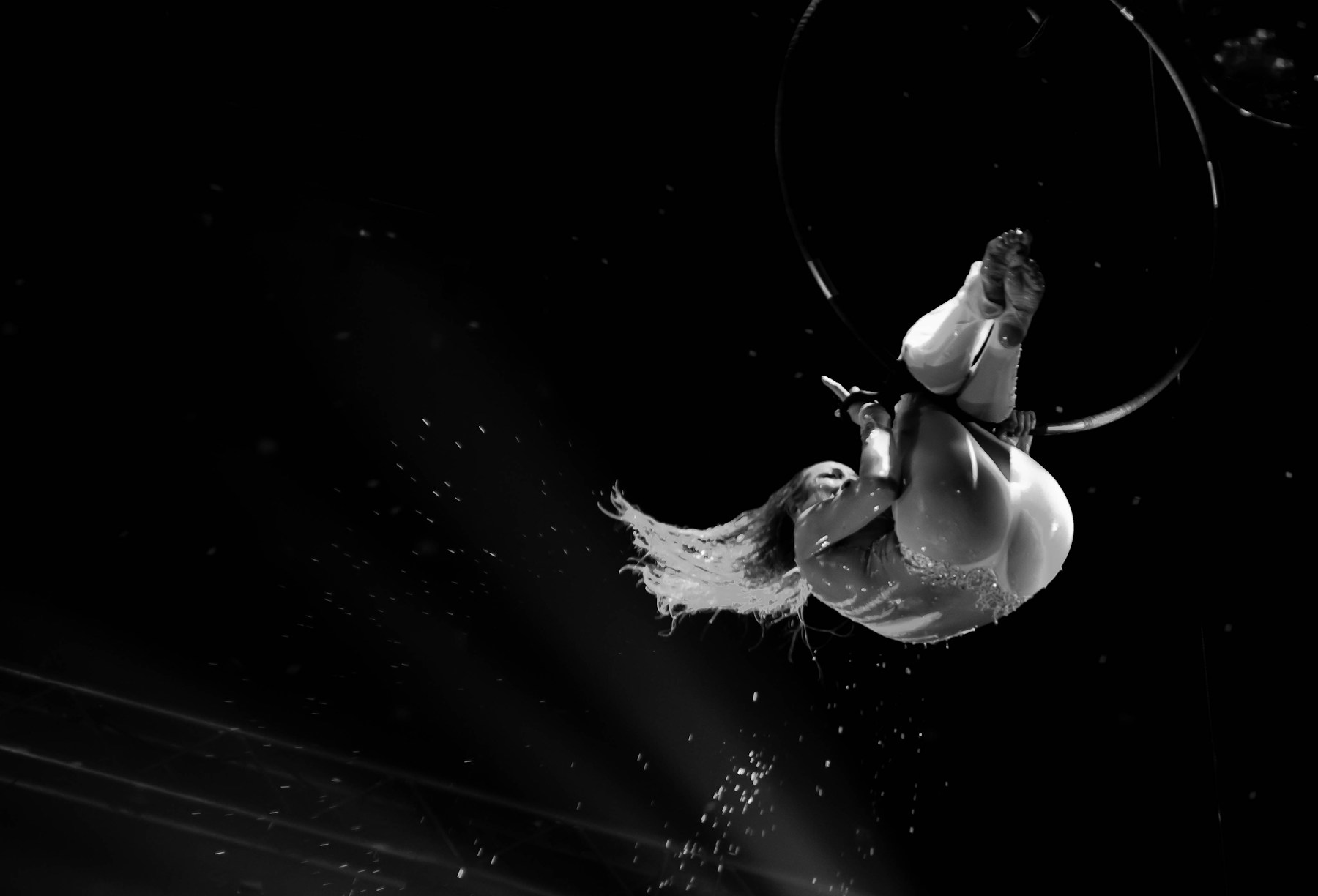 An aerial performer hangs upside down from a hoop against a dark background
