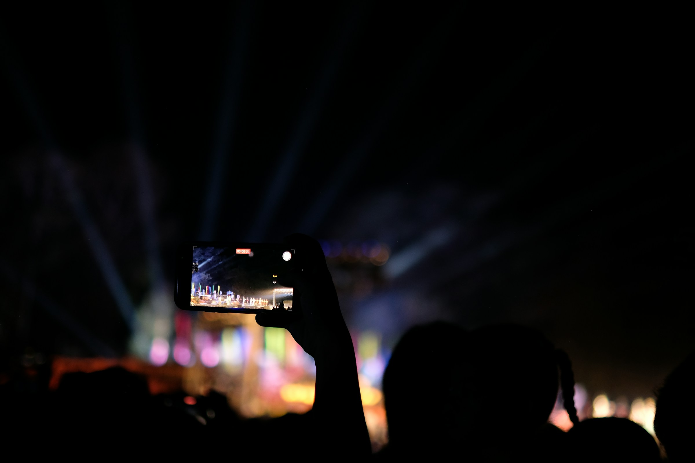A concertgoer records a brightly lit stage on a smartphone in a dark crowd