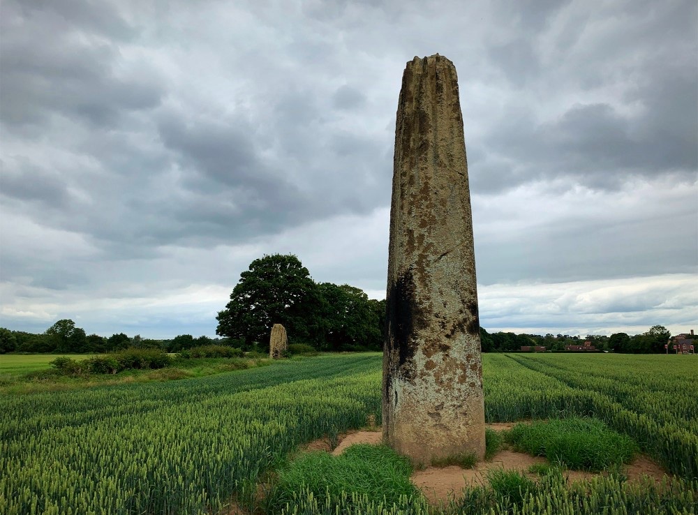 One tall weathered Devil’s Arrow stands in a green field beneath gray clouds, with a second stone visible in the distance