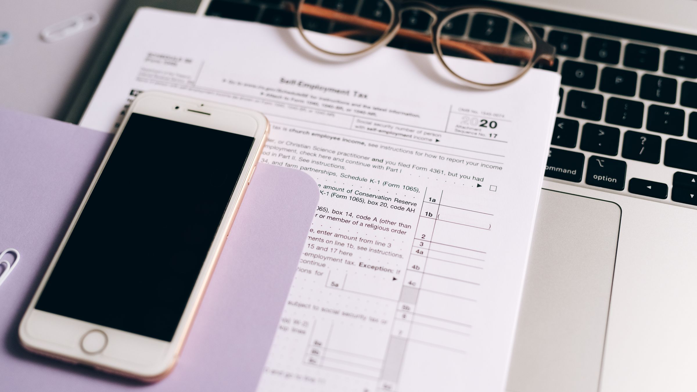 A smartphone, laptop, eyeglasses, and tax documents arranged on a desk