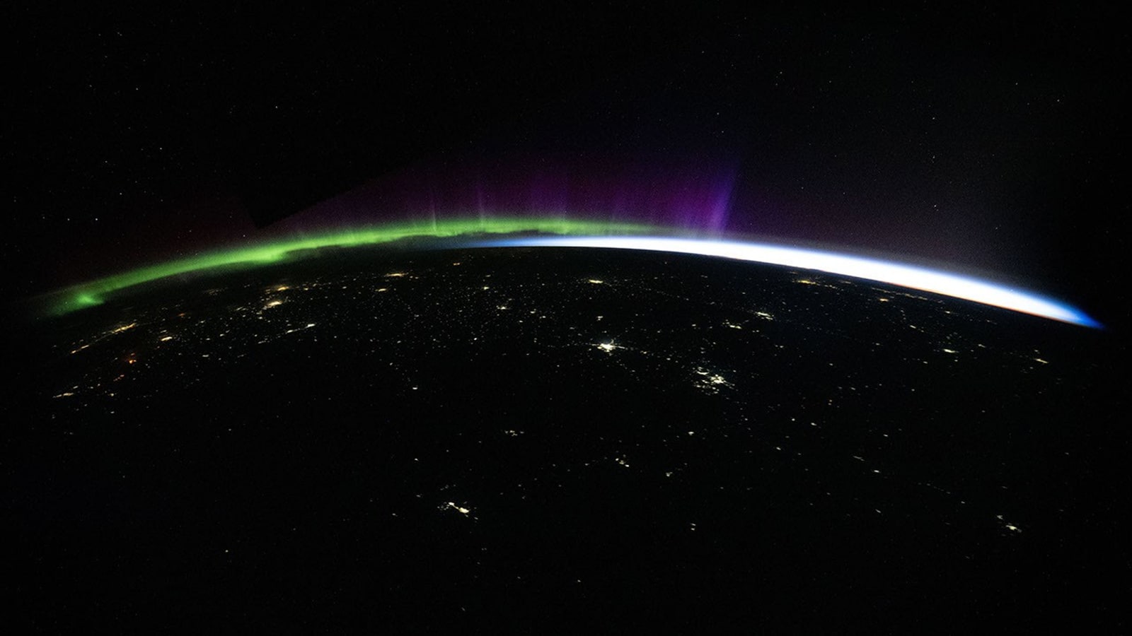 Green and purple aurora rising above Earth's curved night horizon, with city lights scattered across Central Asia below