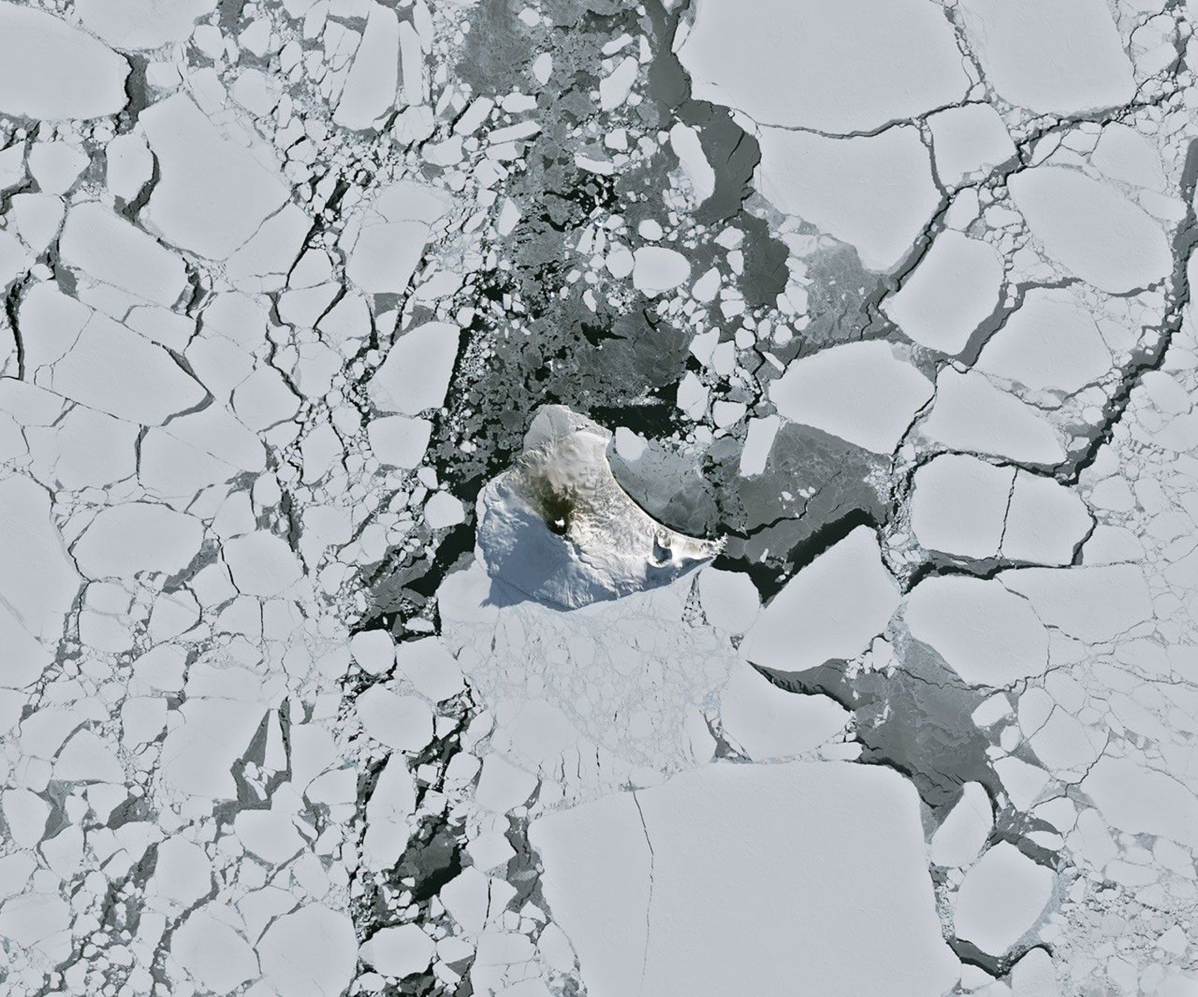 Satellite view of a snow-covered South Atlantic island with fractured sea ice around it and a small red infrared signal at the volcanic summit